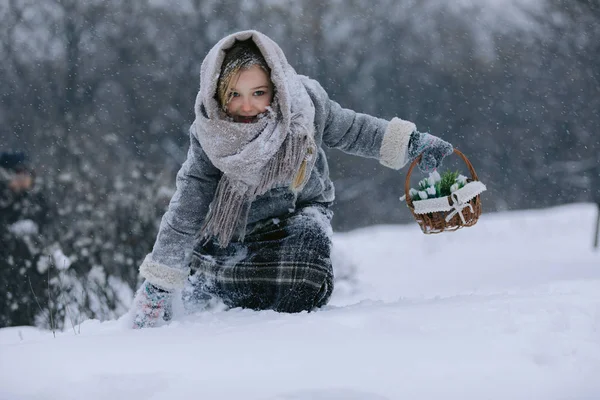 Menina com cesta vai e procura as primeiras flores sob a neve na floresta no inverno — Fotografia de Stock