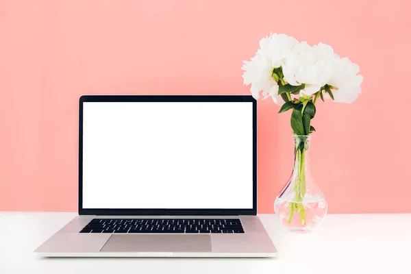 Laptop mit weißem leeren Bildschirm und Blumen in einer Vase auf einem Tisch auf korallenfarbenem Hintergrund. Attrappe — Stockfoto