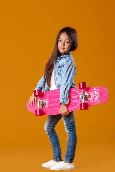 Snygg liten barn flicka med skateboard i denim på orange bakgrund — Stockfoto