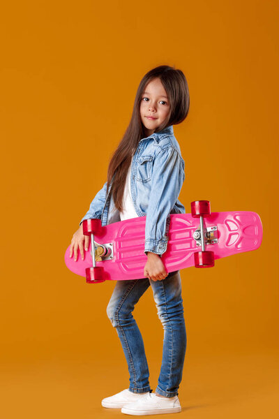 Stylish little child girl with skateboard in denim on orange background