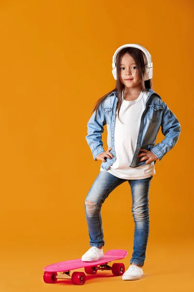 Стильна маленька дівчинка з скейтбордом в денімі на помаранчевому фоні — стокове фото