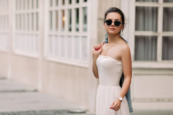 Vonzó nő napszemüveg és fehér ruhában sétál át a város utcáin egy napsütéses napon — Stock Fotó