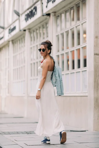 Привлекательная женщина в солнечных очках и белом платье прогуливается по улицам города в солнечный день — стоковое фото