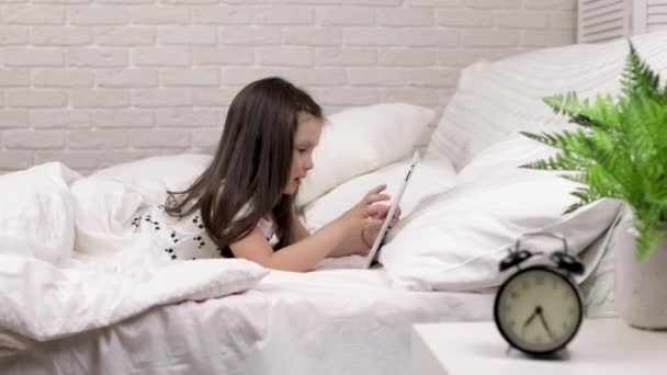 Χαριτωμένο μικρό παιδί κορίτσι βρίσκεται στο κρεβάτι χρησιμοποιεί ψηφιακό δισκίο. — Αρχείο Βίντεο