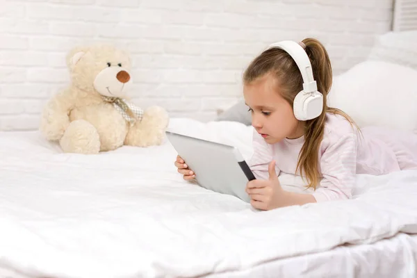 Linda niña se encuentra en la cama utiliza tableta digital . — Foto de Stock