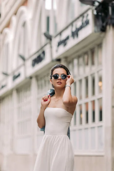 Mulher atraente em óculos de sol e vestido branco caminha pelas ruas da cidade em um dia ensolarado — Fotografia de Stock