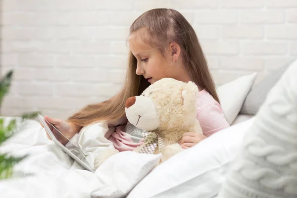 Linda niña se encuentra en la cama utiliza tableta digital . —  Fotos de Stock