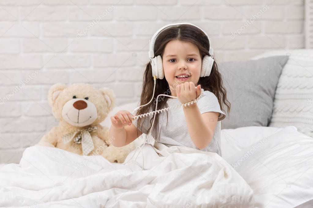 little girl listening to the music with the headphones on bed.