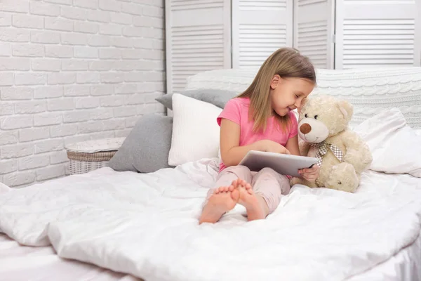 Bonito menina encontra-se na cama usa tablet digital . — Fotografia de Stock