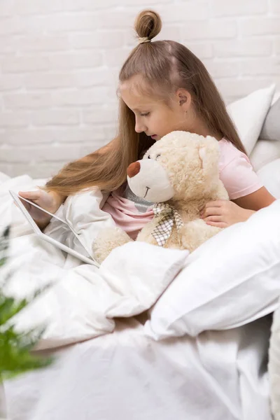 Linda niña se encuentra en la cama utiliza tableta digital . —  Fotos de Stock