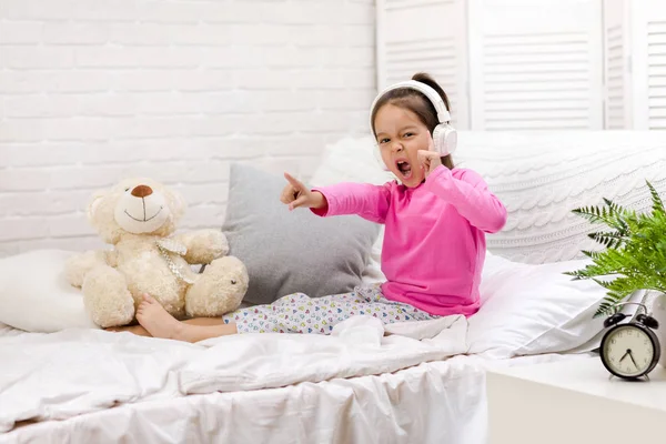 Klein meisje luisteren naar de muziek met de koptelefoon op bed. — Stockfoto