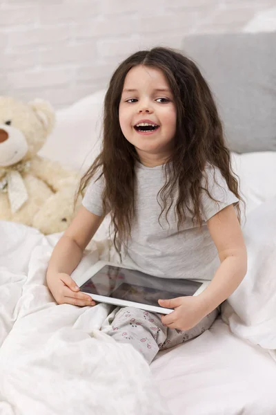 Mignonne petite fille se trouve dans le lit utilise tablette numérique . — Photo