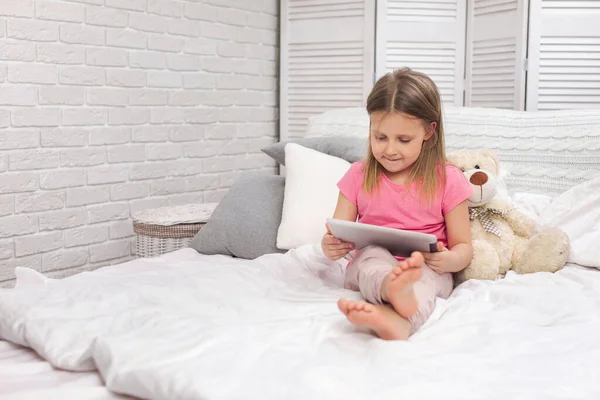 Bonito menina encontra-se na cama usa tablet digital . — Fotografia de Stock