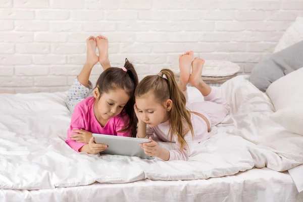 Két aranyos gyerekek lányok használata digitális tabletta az ágyban. — Stock Fotó