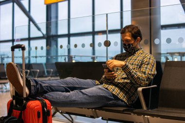 Havaalanında tişört giyen genç ve çekici bir adamın fotoğrafı intihara hazır bir şekilde uçağa yetişmek için bekliyor. Telefonunu kontrol ediyor ve yeniden kullanılabilir yüz maskesi takıyor. Akıllı günlük biçim