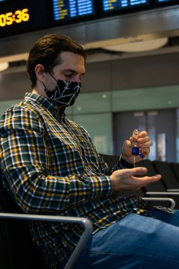 Havaalanında uçak beklerken el dezenfektanı kullanan genç ve çekici bir adamın fotoğrafı. Yeniden kullanılabilir yüz maskesi takıyor.,