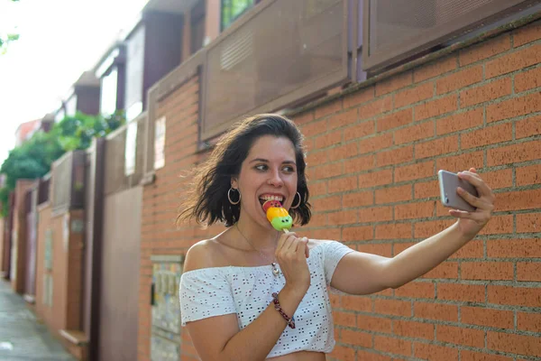 一个年轻貌美的女人一边吃着冰淇淋一边拿着带有橙色背景的手机自拍 — 图库照片