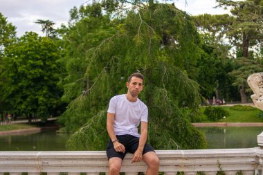 Yazın, basit ve modern giysileri olan, yeniden kullanılabilir yüz maskesi takan ve parkta dikilen genç ve çekici bir adamın fotoğrafı.