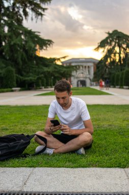 Beyaz tişörtlü, çimlerin üzerinde oturan ve telefonunu kontrol eden genç ve çekici bir adamın fotoğrafı.