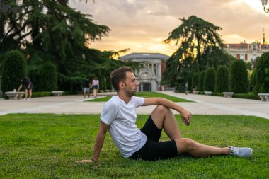 Genç ve çekici bir adamın fotoğrafı. Çimlerin üzerinde rahatça oturup ufka bakıyor.