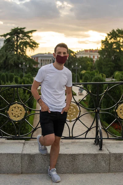 Photo of a young and attractive man wearing a white t-shirt wearing a reusable face mask and checking his phone in the park