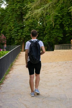 Tekrar kullanılabilir yüz maskesi takan sırt çantalı genç ve çekici bir adamın parkta turizm yaparken çekilmiş fotoğrafı.