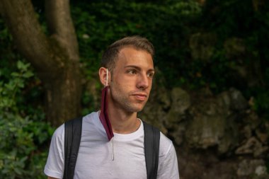 Genç ve çekici bir adamın fotoğrafı. Sırt çantalı, kulağında yeniden kullanılabilir yüz maskesi asılı.. 