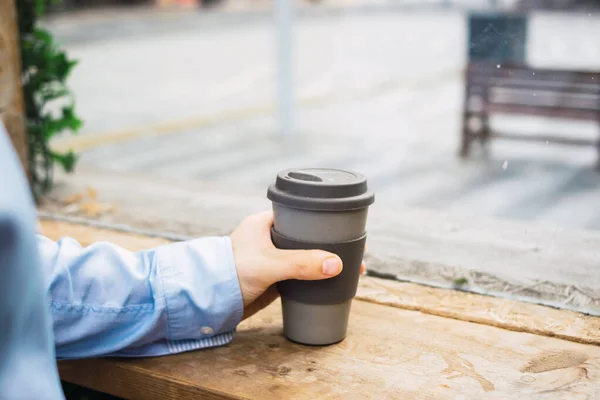 Photo of a reusable cup of coffee on a wood table with the phone. A hand with casual clothes is holding the cup. Job break. Smart working