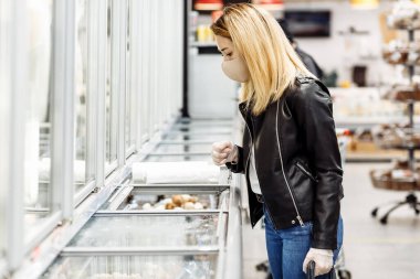 Coronavirus Covid-19 salgını sırasında alışveriş. Genç bir kadın süpermarketten alışveriş arabasıyla yiyecek alır. Enfeksiyonu önlemek için maske ve eldiven takan kadın..