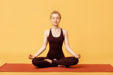 Genç sarışın kadın meditasyon yapıyor, yoga lotusu poz veriyor ve Asana yapıyor. Spor kıyafetleriyle yoga yapan sporcu kız sarı arka planda, izole edilmiş. Stressiz Yojik Meditasyon Uygulaması