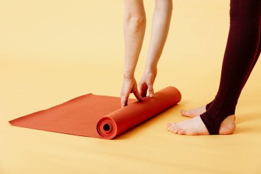Sarı arka planda çalıştıktan sonra kırmızı yoga ya da spor paspası katlayan çekici genç bir kadın. Yoga ve sağlıklı yaşam tarzı konsepti. Yatay çekim. Stüdyo çekimi