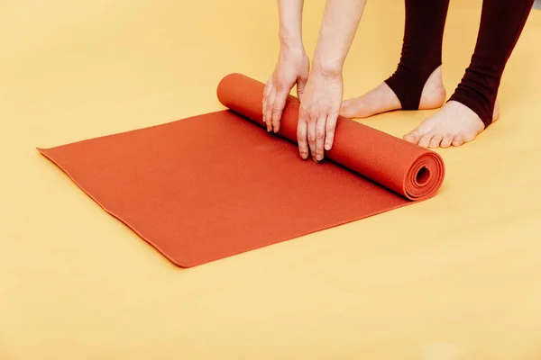 Sarı arka planda çalıştıktan sonra kırmızı yoga ya da spor paspası katlayan çekici genç bir kadın. Yoga ve sağlıklı yaşam tarzı konsepti. Yatay çekim. Stüdyo çekimi