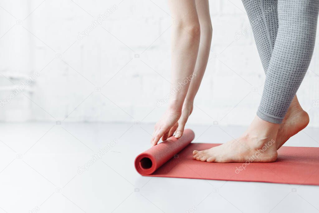 Young yoga Woman rolling her pink mat after a yoga class on wooden floor near a window, close up