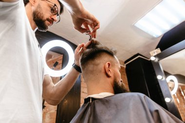 Genç yakışıklı berber berber dükkanında çekici bir adamın saçını kesiyor. Berber dükkanı konsepti. Yakın plan, usta kuaför makas ve tarak ile saç stili ve stili yapıyor..