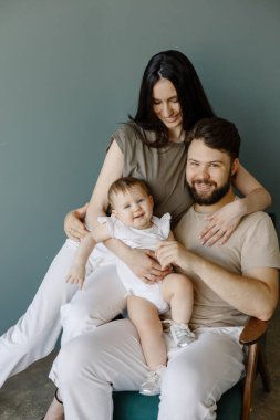 Baba, anne ve küçük kızları mavi arka planda, stüdyoda tahta sandalyede oturuyorlar. Mutlu bir aile, yaşam tarzı, ebeveynler.