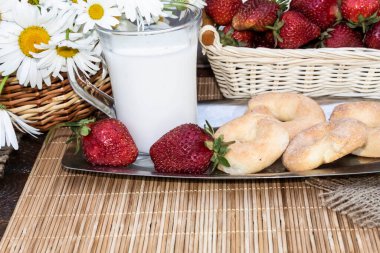 Tepside süt kremalı bir fincan var, yanında da çilek ve kurabiye. Arka planda çilekli bir sepet ve papatyalı bir sepet var.