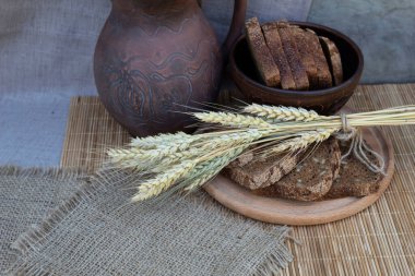 Yuvarlak bir kesme tahtasının üzerinde siyah dilimlenmiş ekmek ve buğday kulakları uzanıyor. Arka planda bir kase ekmek ve bir sürahi var.