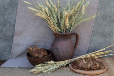 Yuvarlak bir kesme tahtasının üzerinde siyah dilimlenmiş ekmek ve buğday kulakları uzanıyor. Arka planda bir kase ekmek ve buğday kulaklı bir sürahi var..