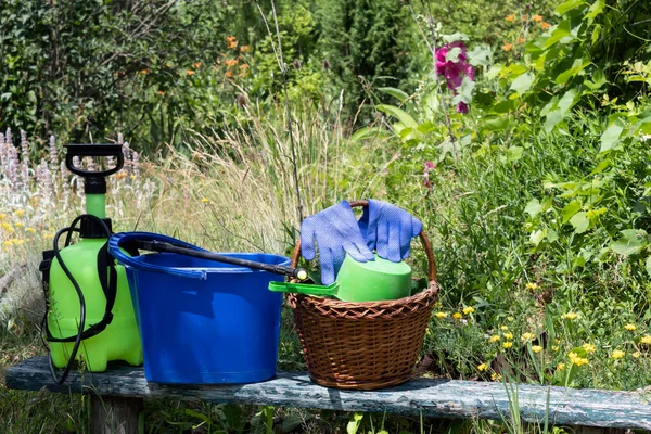 On the garden bench is a bucket of water, a sprayer to combat plant diseases and a basket in which gloves and a mug for water lie.