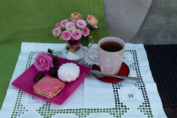 On the table on a white napkin is a bouquet of pink roses, a cup of tea, a plate on which marshmallows, a cake, two candies are lying.