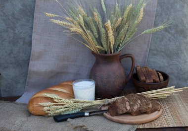 Masanın üzerinde beyaz ekmek yatıyor, bir bardak süt duruyor, bir bıçak ve dilimlenmiş kahverengi ekmek yatıyor. Arka planda buğday kulaklı bir sürahi var..