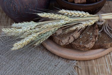 Yuvarlak bir tahtanın üzerinde dilimlenmiş siyah ekmek ve buğday başakları var..