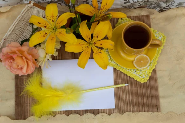 Yukarıdan bak. Masanın üzerinde limonlu bir fincan çay, bir buket sarı zambak ve bir gül var. Üzerinde sarı tüylü bir kalem bulunan bir kağıt yaprağı.