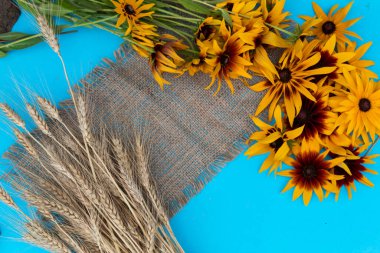 Mavi bir arka planda jüt kumaş üzerinde buğday ve rudbeckia çiçekleri vardır..