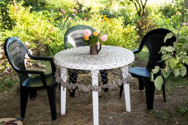 Kırsal bahçede güzel örülmüş bir masa örtüsüyle kaplı beyaz bir masa var. Masanın üzerinde bir buket gül var. Masanın yanında sandalyeler var..