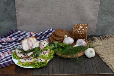 Marul yapraklarının üzerindeki beyaz tabakta pastırma, biber, dereotu ve maydanoz var. Siyah ekmek, sarımsak ve otlar yuvarlak bir tahtanın üzerinde yatıyor.