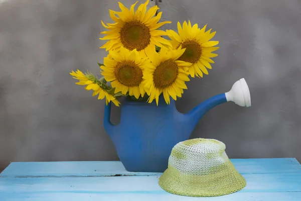 A watering pot with a bouquet of sunflower stands on a blurred background. Nearby lies a summer women\'s hat.