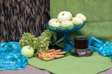 Masadaki bardak tutacağında bir bardak çay var. Yakınlarda bir tabak kurabiye ve şekerli meyve, bir vazo elma, ortanca ve bir elma var..