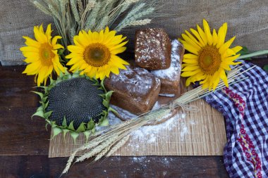 Ahşap bir arka planda, üç somun ekmek, un, buğday kulakları, çiçekler ve ekmek pişirmek için olgun tohumlarla dolu bir ayçiçeği bulunur.