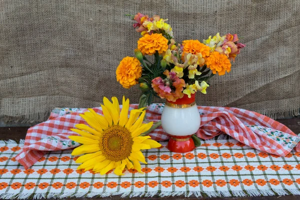 Against the background of jute fabric, on a colored napkin in a vase, there is a bright bouquet of tagetis and snapdragon flowers. Nearby lies a sunflower flower